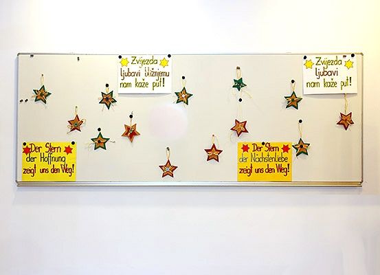 Ein weißes Board mit verschiedenen sternförmigen Dekorationen. Einige Sterne haben deutschen Text, während andere englisch sind. Der deutsche Text lautet, 'Der Stern der Hoffnung zeigt uns den Weg!' und 'Der Stern der Nachslenliebe zeigt uns den Weg!' Der englische Text lautet, 'The star of hope shows us the way!' und 'The star of love shows us the way!'.