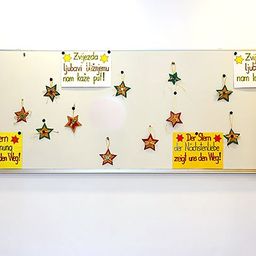 Ein weißes Board mit verschiedenen sternförmigen Dekorationen. Einige Sterne haben deutschen Text, während andere englisch sind. Der deutsche Text lautet, 'Der Stern der Hoffnung zeigt uns den Weg!' und 'Der Stern der Nachslenliebe zeigt uns den Weg!' Der englische Text lautet, 'The star of hope shows us the way!' und 'The star of love shows us the way!'.