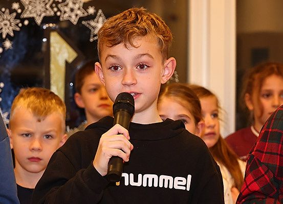 Ein junger Junge in einem schwarzen HUMMEL-Sweatshirt spricht in ein Mikrofon, umgeben von Kindern.
