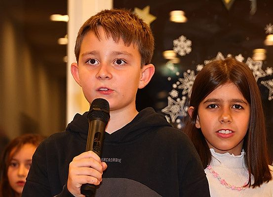 Ein Junge und ein Mädchen stehen vor einer festlichen Wand mit Schneeflocken und halten ein Mikrofon. Der Junge spricht, während das Mädchen aufmerksam zuhört. Hinter ihnen steht ein weiteres Mädchen, das ebenfalls zuhört.