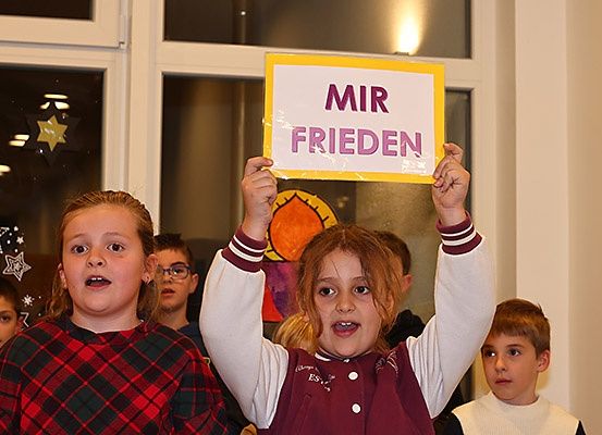 Ein junges Mädchen hält ein Schild mit den Worten 'MIR FRIEDEN' vor anderen Kindern in einem Raum mit einem dekorierten Fenster.