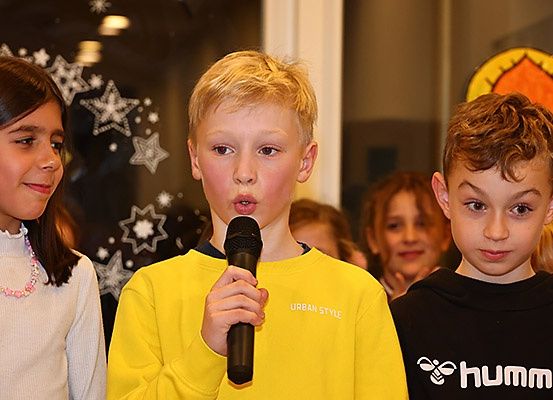 Drei Kinder stehen vor einer Menschenmenge, ein Junge in Gelb hält ein Mikrofon und spricht. Ein Mädchen in Weiß und ein Junge in Schwarz stehen neben ihm. Der Hintergrund ist mit Sternen und einem gelben Kreis verziert.