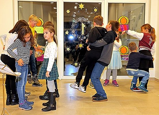 Eine Gruppe von Kindern spielt und tanzt in einem Raum mit dekorierten Glastüren und einem Notenständer.
