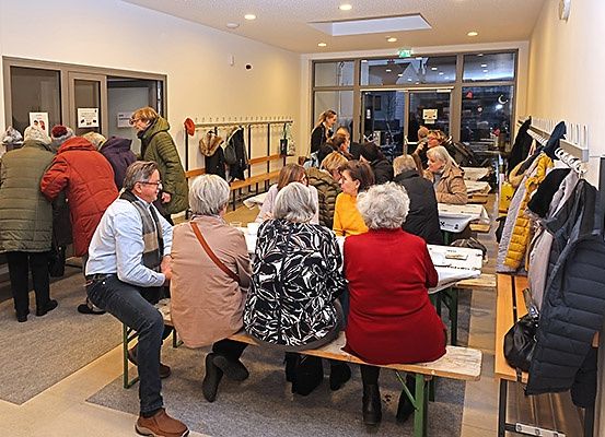 Eine Gruppe von Menschen in Winterkleidung sitzt und steht an Tischen in einem Raum mit großen Fenstern und Kleiderbügeln.