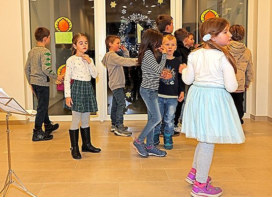 Eine Gruppe von Kindern steht in einem Raum, einige singen und einer hält einen Notenständer. Sie sind in Winterkleidung gekleidet.