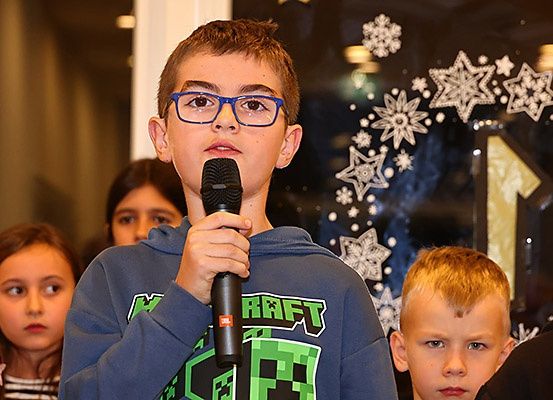 Ein junger Junge mit Brille hält ein Mikrofon und spricht zu einer Gruppe von Kindern in einer festlichen Umgebung.