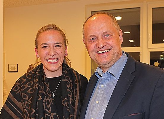 Ein Mann und eine Frau stehen eng beieinander und lächeln für ein Foto. Die Frau trägt einen gemusterten Schal und eine Halskette, während der Mann einen dunklen Anzug und ein hellblaues Hemd trägt. Hinter ihnen ist eine Wand mit einem Schild und zwei Fenstern zu sehen.