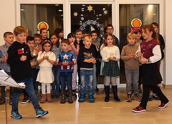 Eine Gruppe von Kindern steht in einer Reihe, hält Tassen und scheint zu singen. Sie befinden sich in einem Raum mit Glastüren, der mit Sternen dekoriert ist.