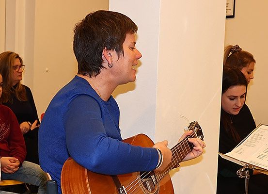 Eine Frau spielt Gitarre, während eine Gruppe von Menschen vor ihr sitzt, einige hören aufmerksam zu, andere lesen aus einem Notenblatt.