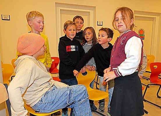 Eine Gruppe von Kindern ist in einem Klassenzimmer. Einige sitzen auf gelben Stühlen, andere stehen herum. Ein Mädchen in einer kastanienbraunen Jacke führt die Gruppe an.