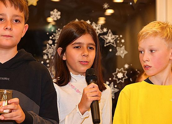 Drei Kinder stehen eng beieinander, eines hält eine Glaskerze und ein anderes ein Mikrofon.