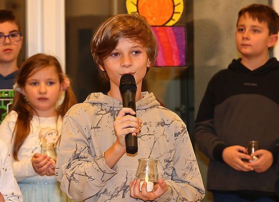 Ein junger Junge mit einem Mikrofon steht vor einer Gruppe von Kindern mit Kerzen, wahrscheinlich während einer Veranstaltung.
