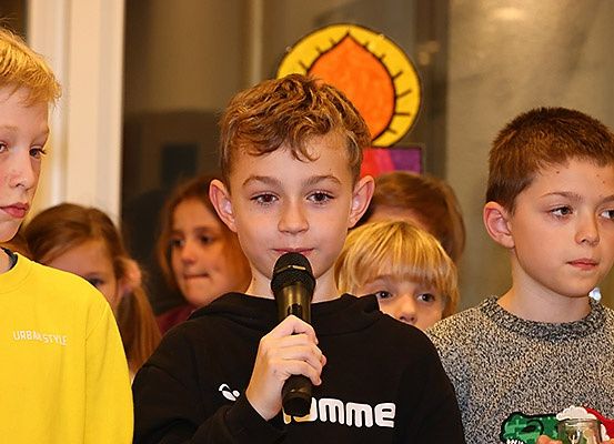 Eine Gruppe von Kindern steht in einem Raum, von denen einer ein Mikrofon in der Hand hält. Der Junge in der Mitte schaut nach vorne, während er in das Mikrofon spricht. Der Junge auf der linken Seite trägt ein gelbes T-Shirt. Hinter ihnen stehen weitere Kinder. Eine Lampe ist an der Wand montiert.