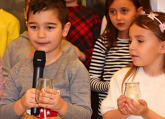 Ein kleiner Junge hält ein Mikrofon und eine Kerze in der Hand und steht neben einem Mädchen mit einer Kerze. Sie sind von anderen Kindern umgeben.