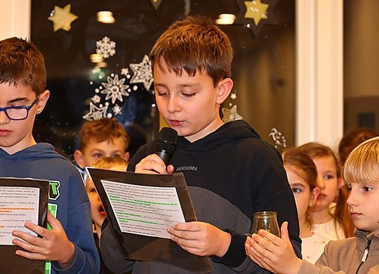 Mehrere junge Kinder stehen zusammen und halten Papiere und ein Mikrofon. Ein Junge spricht in das Mikrofon, während andere ihn aufmerksam beobachten. Im Hintergrund befindet sich ein Glasfenster mit dekorativen Schneeflocken und Sternen.