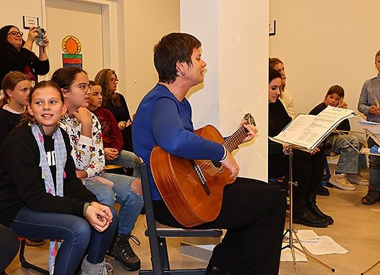 Eine Frau spielt Gitarre, während eine Gruppe von Kindern und Erwachsenen aufmerksam zuhört. Die Umgebung scheint ein Klassenzimmer oder eine ähnliche Bildungsumgebung zu sein.