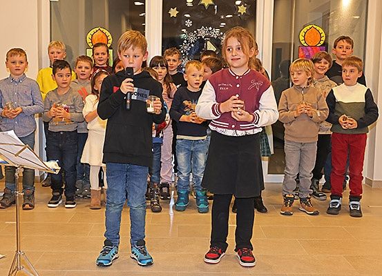 Kinder, die Kerzen halten, stehen in einer Reihe in einem Raum, zwei halten Mikrofone in der Front. Sie scheinen zu singen.