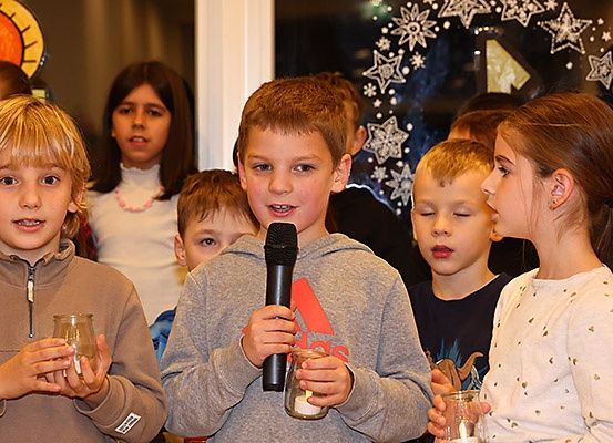 Eine Gruppe von Kindern steht zusammen und hält Kerzen, wobei ein Junge ein Mikrofon hält und spricht. Sie sind in einem Raum versammelt, möglicherweise für eine Zeremonie.