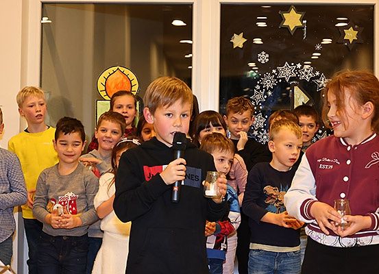 Eine Gruppe von Kindern versammelt sich drinnen, einer hält ein Mikrofon, wahrscheinlich während einer Feiertagsveranstaltung. Hinter ihnen schmücken Dekorationen die Glasfenster.