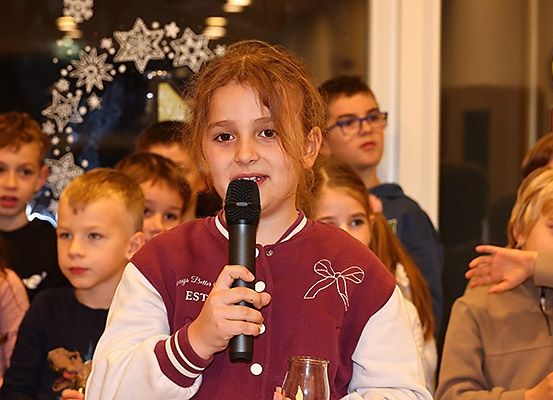 Ein Mädchen mit roten Haaren steht vor einer Gruppe von Kindern, hält ein Mikrofon und einen Glas in der Hand. Schneeflocken schmücken den Hintergrund.