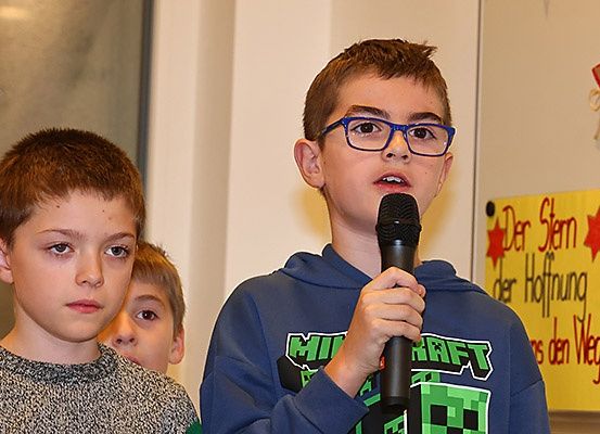 Drei Jungen stehen in einem Raum. Der Junge in der Mitte spricht in ein Mikrofon, während der Junge auf der linken Seite ihn ansieht. Dahinter befindet sich ein Glasfenster.
