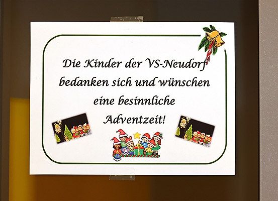 Ein weißes Schild mit grünem Rand zeigt eine Weihnachtsbotschaft auf Deutsch. Es enthält eine Illustration von Kindern in Weihnachtsmützen, einer Glocke und Weihnachtsbäumen. Die Botschaft übersetzt sich zu 'Die Kinder der VS-Neudorf danken und wünschen eine besinnliche Adventszeit!'