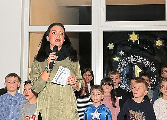 Eine Frau mit Mikrofon spricht eine Gruppe von Kindern an, die vor einem Fenster mit Dekorationen stehen.