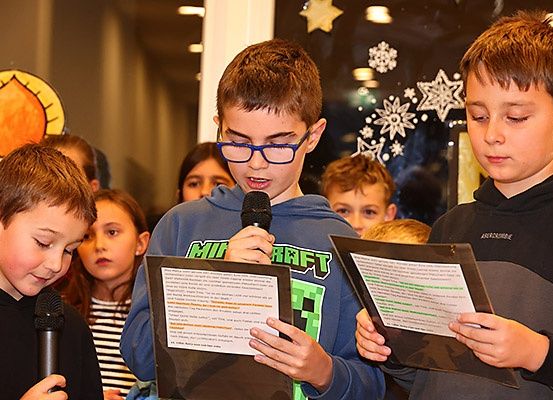 Drei Jungen halten Papiere und Mikrofone in der Hand und lesen wahrscheinlich aus einem Skript in einem Klassenzimmer. Hinter ihnen schaut eine vielfältige Gruppe von Kindern aufmerksam zu. Eine Weihnachtsdekoration ist an der Wand zu sehen.