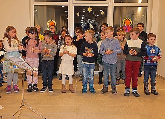 Mehrere Kinder stehen in einer Reihe und singen in einem Raum mit großen Fenstern, einige halten kleine Kerzen. Eine Person hält einen Notenständer.