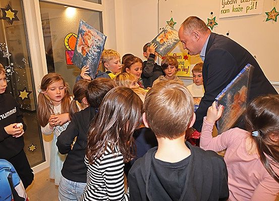 Ein Mann in einem Anzug verteilt Geschenke an eine Gruppe von Kindern, die sich um ihn versammelt haben. Die Kinder halten Geschenke und schauen den Mann an.