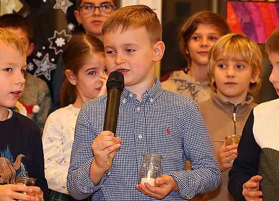 Eine Gruppe von Kindern, einer mit einer Kerze, steht im Kreis. Ein Junge mit Brille und Mikrofon spricht zu ihnen.