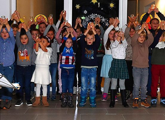 Eine Gruppe von Kindern, in Winterkleidung gekleidet, steht zusammen und hält brennende Kerzen. Einige tragen Stiefel, andere Sneakers. Links steht ein Notenständer, und eine Wand mit Sternen und Schneeflocken ist im Hintergrund.