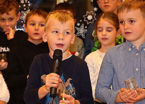 Eine Gruppe von Kindern ist versammelt, einige halten Kerzen. Ein Junge spricht in ein Mikrofon, während andere zuhören. Der Hintergrund zeigt festliche Dekorationen.