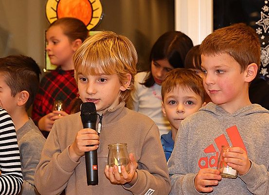 Eine Gruppe von Kindern, die Kerzen halten, steht in einem Raum. Ein Junge in einem braunen Hoodie hält ein Mikrofon und eine Glaskerze, während ein anderer Junge in einem grauen Hoodie eine Glaskerze hält. Hinter ihnen stehen andere Kinder und schauen zu.