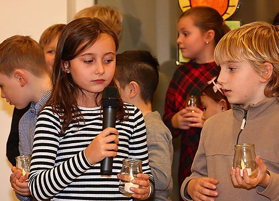 Eine Gruppe von Kindern in einem Raum, einige halten Kerzen. Ein Mädchen mit einem Mikrofon scheint zu sprechen. Ein Junge neben ihr hält eine Kerze in der Hand.