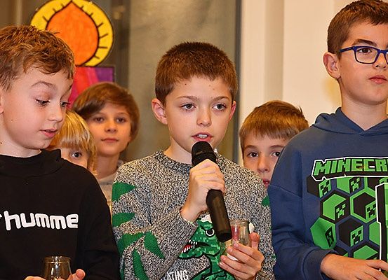 Eine Gruppe von Jungen steht in einem Raum, einer hält ein Mikrofon und ein anderer ein Glas. Im Hintergrund befindet sich eine dekorative Lampe.