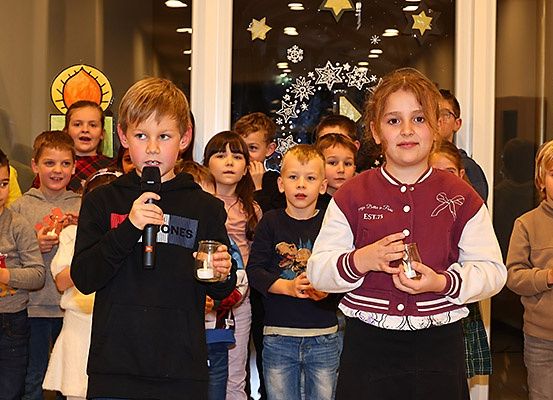 Mehrere Kinder stehen in einer Reihe und halten Kerzen. Ein Junge spricht in ein Mikrofon, während ein Mädchen eine Kerze hält. Dahinter schauen andere Kinder zu, und Erwachsene stehen in der Nähe eines Fensters mit Dekorationen.