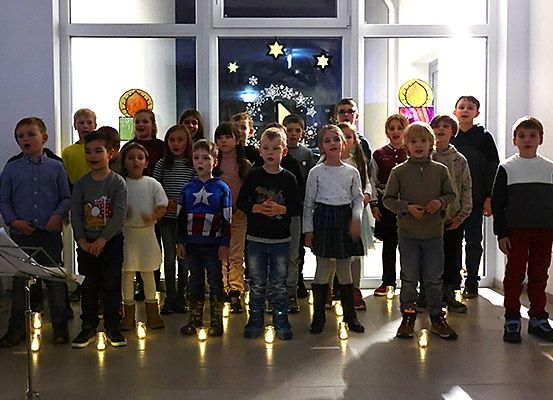 Eine Gruppe von Kindern in verschiedenen Outfits steht in einer Reihe, singt mit offenem Mund und wird von Kerzen beleuchtet. Hinter ihnen ist eine Glastür mit Sternendekoration.