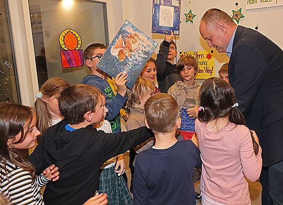 Ein Mann in einem Anzug verteilt Geschenke an eine Gruppe von Kindern in einem Klassenzimmer. Einige Kinder halten Geschenke, und ein Kind hält ein Buch.