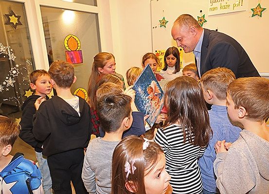 Ein Mann liest ein Buch für eine Gruppe von Kindern in einem Klassenzimmer vor. Die Kinder hören aufmerksam zu, und einige von ihnen halten das Buch in den Händen. Das Klassenzimmer hat Glasfenster mit Zeichnungen darauf.