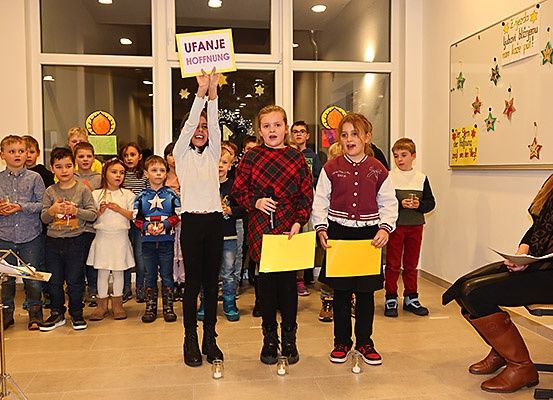 Eine Gruppe von Kindern steht in einer Reihe, hält Schilder und Kerzen und singt vielleicht. Dahinter befindet sich eine Wand mit Sternen und eine Tafel mit Dekorationen.
