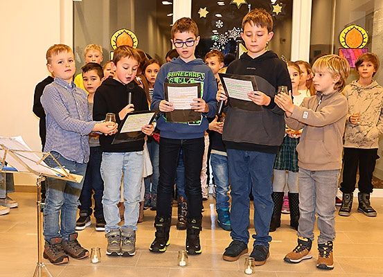 Mehrere Kinder stehen in einem Raum, halten Papiere und Kerzen und nehmen möglicherweise an einer Schulveranstaltung teil. Sie scheinen einen Gesang oder ein Lied aufzuführen.