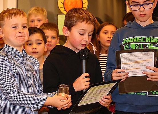 Mehrere Kinder stehen in einem Klassenzimmer, einige halten Papiere und ein Mikrofon. Ein Kind trägt ein Minecraft-Sweatshirt.