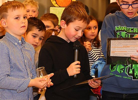Ein junger Junge hält ein Mikrofon in der Hand und liest laut vor im Klassenzimmer. Ein weiterer Junge neben ihm hält einen Glasbecher.