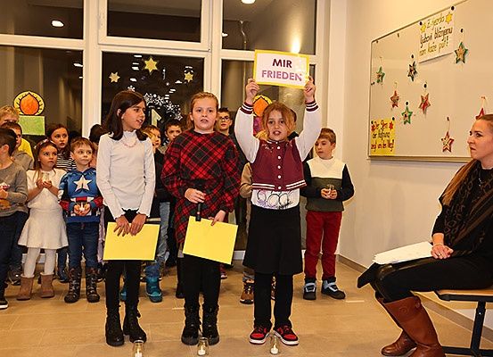 Eine Gruppe von Kindern, die gelbe Papiere und Schilder halten, eines von ihnen hält ein Schild mit der Aufschrift MIR FRIEDEN, stehen in einem Raum mit einer Frau, die auf einer Bank sitzt.