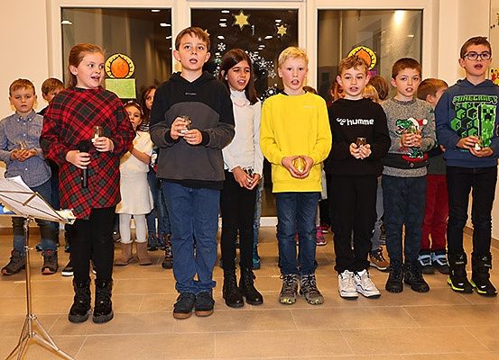 Eine Gruppe von Kindern steht drinnen zusammen, wahrscheinlich bei einer Schulveranstaltung. Sie halten kleine Gegenstände und scheinen zu singen. Einige tragen Brillen und Turnschuhe. Der Raum hat einen gefliesten Boden und Dekorationen an den Wänden.
