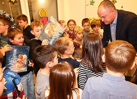 Eine Gruppe von Kindern und ein Mann versammeln sich drinnen, wahrscheinlich in einem Klassenzimmer. Der Mann liest aus einem Buch vor, während die Kinder aufmerksam zuhören, einige halten ihre eigenen Bücher.