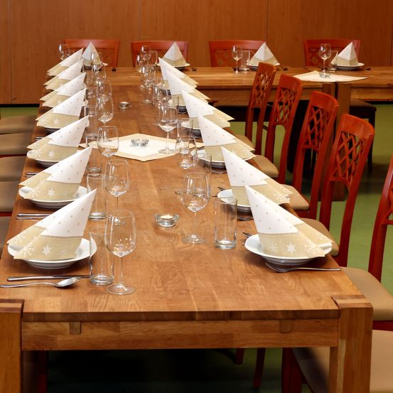 Ein langer Holztisch, der für eine formelle Veranstaltung gedeckt ist, mit mehreren Platzeinstellungen, jede mit einer weißen Serviette, einem Weinkelch und einem Teller. Die Stühle sind rot und um den Tisch herum angeordnet.
