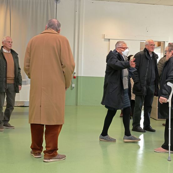 Eine Gruppe älterer Menschen in einem Raum, die Mäntel und Brillen tragen, einer hält einen Stock. Eine Person mit Maske gestikuliert. Ein anderer trägt Turnschuhe. Eine Tafel ist an der Wand.
