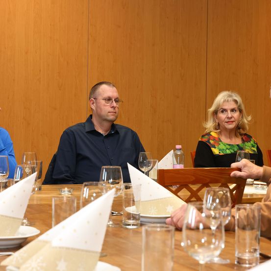 Eine Gruppe von Menschen sitzt um einen Tisch in einem Konferenzraum. Ein Mann und eine Frau sind einander gegenüber, mit Weingläsern, Tellern und Servietten auf dem Tisch. Die Wand hinter ihnen ist aus Holz.